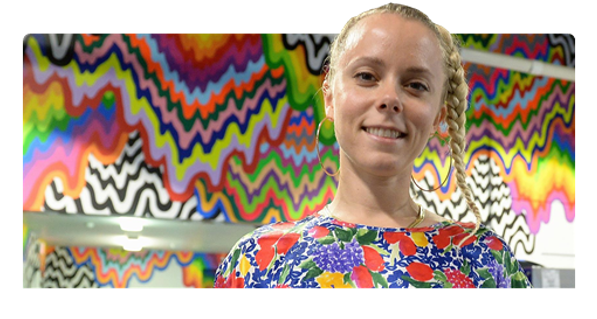 A woman with braided blonde hair smiles in front of a vibrant, multicolored, drip artwork wall. She is wearing a floral top with bright red, purple, and green colors.